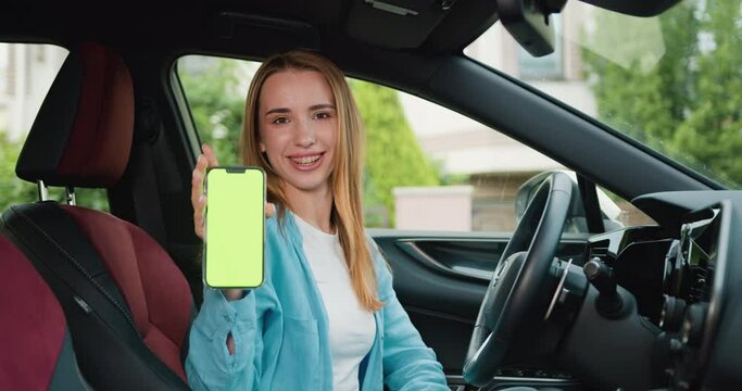Happy smiling woman driver sitting behind the wheel of premium modern car showing smartphone with vertical green screen. Green screen, blank phone, application. Chroma key smartphone technology