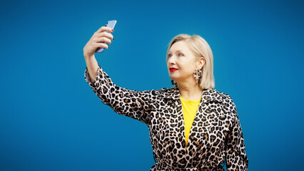 A smiling woman in her 50s is posing for a smartphone camera, taking a selfie for media purposes