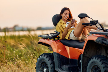 Naklejka premium A smiling woman lying on a rented quad bike and using a mobile phone.