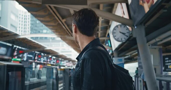 Young man in black shirt with backpack stands on the platform of the ground metro station, waiting for metro train. Caucasian male 30 waits for the train at the subway station public transport