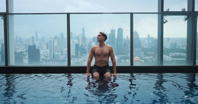 Man chilling and relaxing in luxury pool on roof of skyscraper. Young 30s business male sitting on the edge of the roof top swimming pool in luxury hotel on background big city