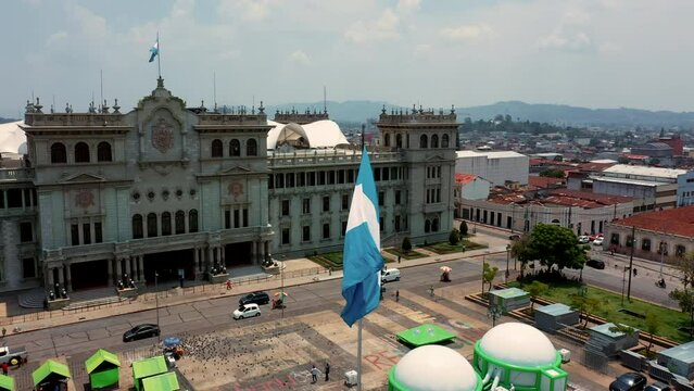 Bandera Guatemalteca ondenado al viento sobre asta en plaza municipal de la ciudad con palacio nacional de cultura de fondo  guatemala y la catedral metropolitana en el centro de la ciudad