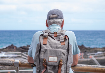 Back view of senior man with cap looking at sea and horizon over water carrying backpack enjoying vacation or travel