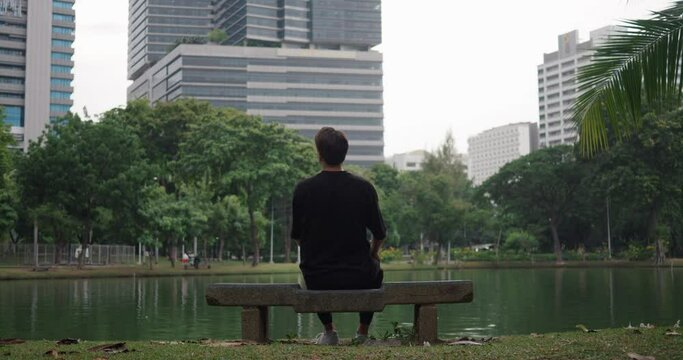 Young 30s man sits on a park bench against the backdrop of a lake in a city park outdoors. Lonely man walks in the park, rests on a bench in the park on backdrop of a lake and buildings in big city