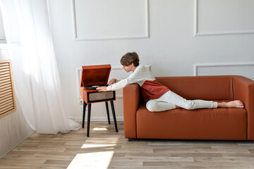 Teenager boy sitting on sofa, turning on wooden vinyl player. Retro aesthetic, indoor, home interior