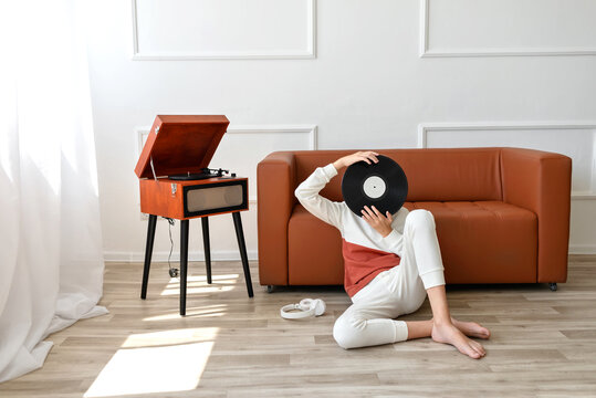 Vinyl record modern trend, youth culture. Teenager boy sitting on floor near sofa and turntable, hiding face behind vinyl record. Lifestyle home minimalist interior
