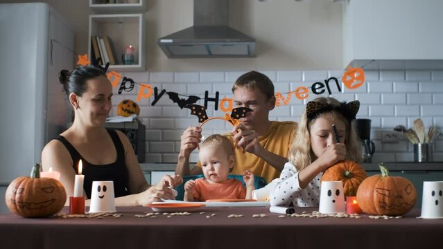 Cheerful family preparing for Halloween party. Man and woman trying on headband with bat wings on her baby while older daughter painting scary face on pumpkin