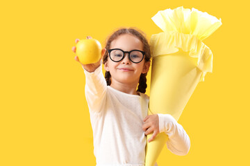 Happy little girl in eyeglasses with school cone and fresh apple on yellow background