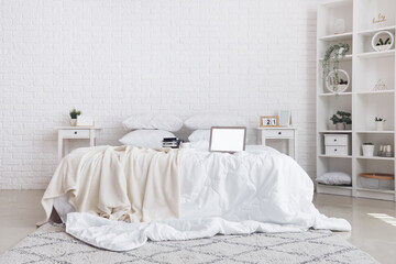 Messy bed with white blanket, coffee on tray and modern laptop in light bedroom