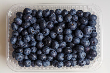 Fresh juicy blueberries in a transparent plastic container with a slide on a white table.