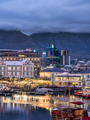 Fototapeta na wymiar V and A Waterfront lit up with the table mountain in the background during a cloudy evening, Cape Town, South Africa