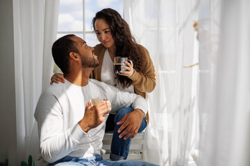 Smiling young asian woman hugging african american boyfriend with coffee on windowsill at home in morning