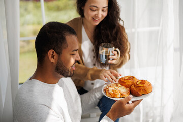 Smiling young african american man holding homemade pastry near asian girlfriend with coffee at home