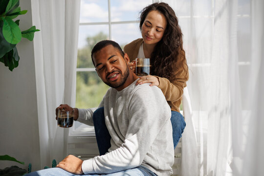 Smiling interracial couple holding cups of coffee while sitting and relaxing near window in morning