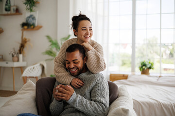 Smiling young asian woman in sweater hugging african american boyfriend holding coffee at home
