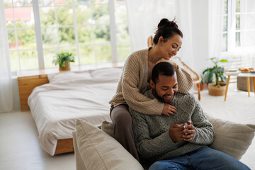 Young asian woman in warm clothes hugging african american boyfriend with coffee on armchair at home