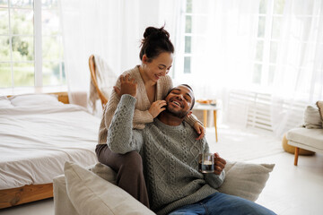 Smiling asian woman hugging african american boyfriend in sweater with coffee on armchair at home