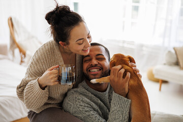 Positive interracial couple in cozy sweaters holding coffee while playing with chihuahua dog at home