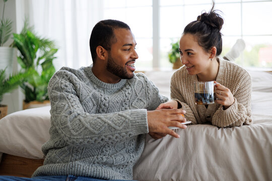 Smiling young multiethnic couple in sweaters talking near coffee and sweet pastry in bedroom at home