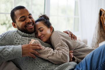 Smiling young asian woman in sweater hugging african american boyfriend while sitting on couch at...