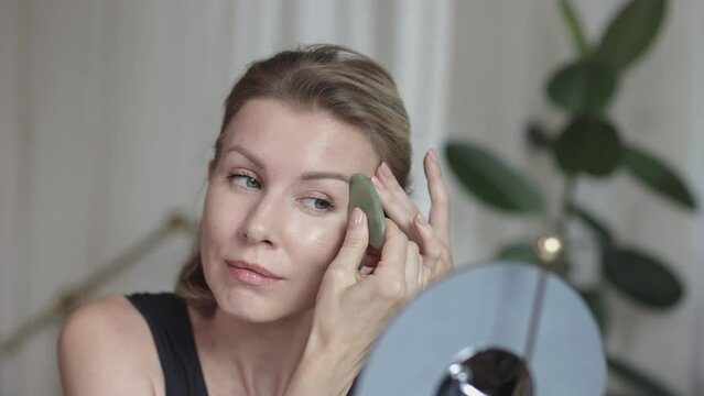Middle aged beautiful woman making herself facial massage with gua sha behind the mirrow at home