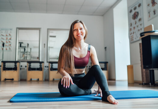 Beautiful fit-shape female dressed sporty sitting and sincerely smiling on sporty gym mat in sport athletic gym. Active people training or medical rehabilitation concept image