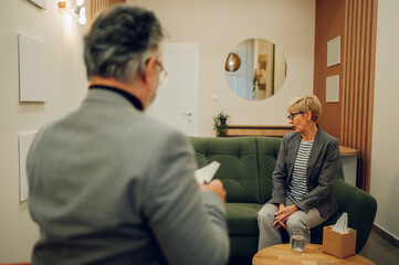 A senior woman with problems is sitting on a couch in a psychotherapist office.