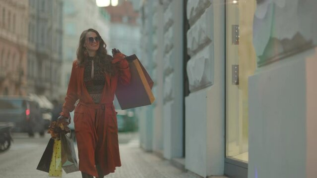 Hello november. happy trendy middle aged woman in brown trench coat with shopping bags and autumn yellow leaves in the city.
