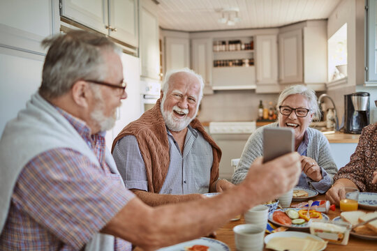 Senior friends using a smartphone while having breakfast together in the kitchen in the morning