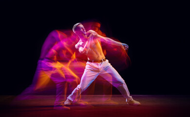 Inspiration and freedom. Young shirtless guy making chaotic movements against black studio background in neon with mixed lights effect. Concept of movements, art, dance and sport, fashion, youth, ad