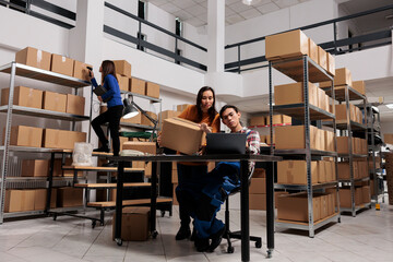 Postal service workers checking parcel invoice on laptop before dispatching while working in storage room. Storehouse employees analyzing freight customs clearance information on computer