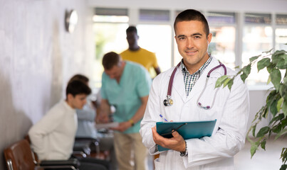 Portrait of smiling polite male doctor in white coat with phonendoscope around neck meeting patient in lobby of clinic, filling out medical form at clipboard - obrazy, fototapety, plakaty