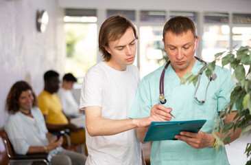 Professional doctor showing papers while explaining results of medical examination to young male patient in lobby of clinic - obrazy, fototapety, plakaty