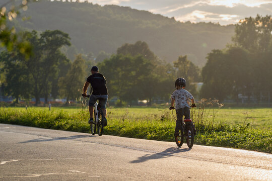A father and his son are riding a bicycle on a sunny evening. They are riding on a path surrounded by greenery and nature. Contrasting sunlight. This is an image of family love and outdoor activities