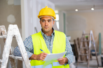 Young foreman in uniform checks the completed construction work and signs documents while standing on the construction site ..indoors