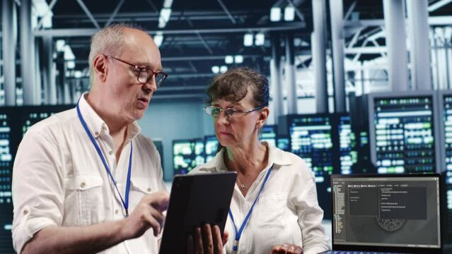 Older coworkers in data center, solving tasks on tablet while running scripts on laptop terminal. Experienced colleagues amidst servers, accessing databases and manipulating lines of code