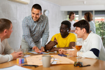 Positive adult man playing board game with interest with male friends of different nationalities during bachelor party