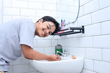 Young Asian man washing hand in the washbasin while looking at camera with happy expression 