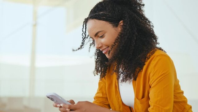 Using gadgets. Close-up side view photo of a pretty, curly haired hispanic or brazilian woman, uses her smart phone, reading news, messaging in social media, texting with family, smile happily
