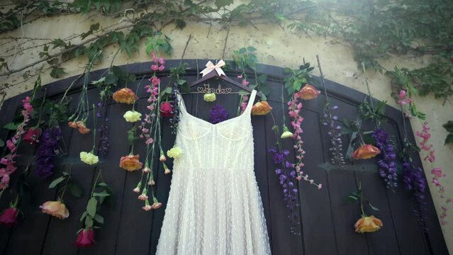 Long white simple wedding dress on a hanger hanging on the black door of the building for wedding ceremony decorated with flowers, hanging upside down. High quality 4k footage