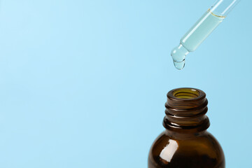 Dripping cosmetic oil from pipette into bottle on light blue background, closeup. Space for text