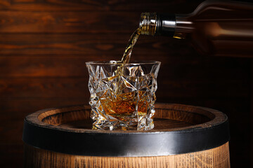 Pouring whiskey into glass from bottle on wooden barrel against brown background