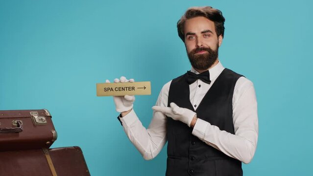 Hotel porter presents direction of spa center using pointer, showing tourists to the right way with wall symbol. Young adult bellhop posing with indicator for relaxation area direction.