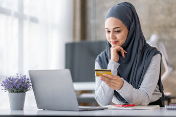 Young beautiful Asian woman in hijab sitting at home office, Muslim woman holding bank credit card...