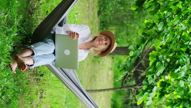 Female freelancer wearing hat using a laptop outdoors. Middle-aged caucasian woman is working relaxed sitting in a hammock among green trees in a summer garden in the backyard.