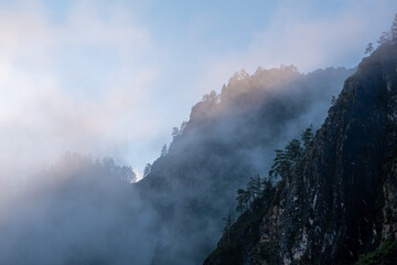 Mountains and peaks in fog at sunrise or sunset. Sun from behind the mountain. Amazing view of mountains and forest landscape with cloudy skies Altai mountains. Mountains and peaks in fog at sunrise