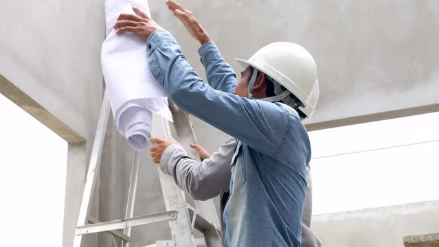 Two male engineers team holding blueprints talking explaining discussing property construction site, Indian architect man with mustache wearing helmet climbing pointing roof structure to be fixed