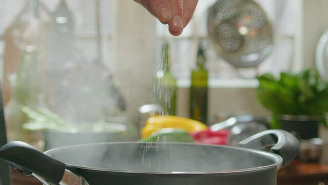 Hand of man adding salt to boiling water in cooking pot, preparing pasta or soup in the kitchen. Close-up shot, selective focus