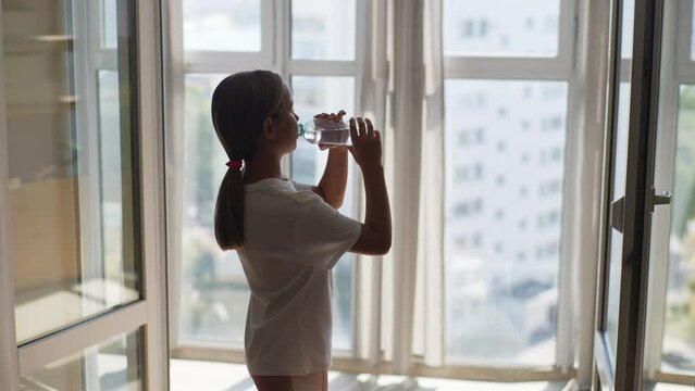 Side view of cute child girl drinking water from plastic bottle preventing dehydration standing by window at home in sunny day. Pretty kid with good life habits, healthy slimming, slow motion.