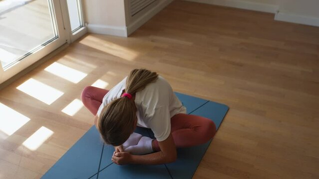 Top view of flexible preteen girl sitting butterfly pose bending forward during yoga home workout., in sunny living room by window. Concept of exercises for children at home and healthy lifestyle
